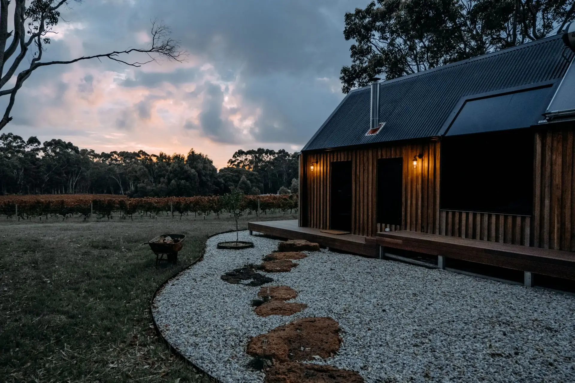 A timber cabin at dusk with its outside lights on, beside a vineyard